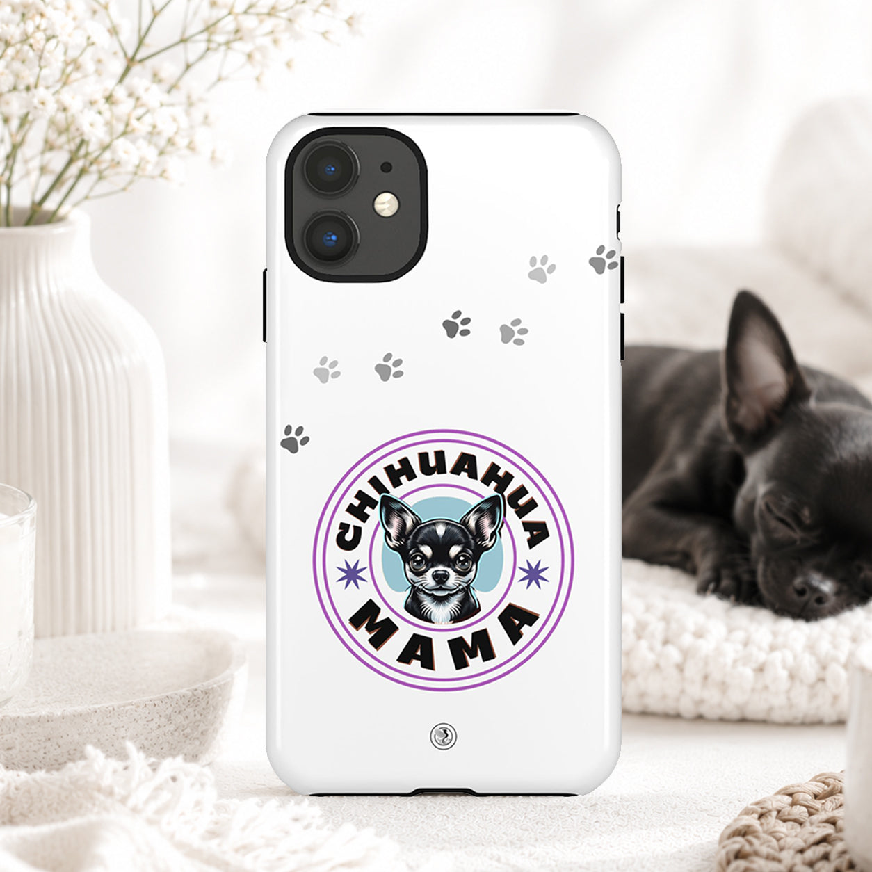 A white phone case featuring a 'Chihuahua Mama' graphic with a chihuahua illustration and paw prints, displayed in a cozy home setting with a sleeping black chihuahua in the background.