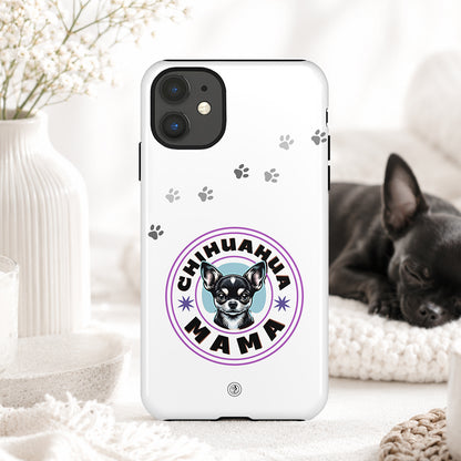 A white phone case featuring a 'Chihuahua Mama' graphic with a chihuahua illustration and paw prints, displayed in a cozy home setting with a sleeping black chihuahua in the background.