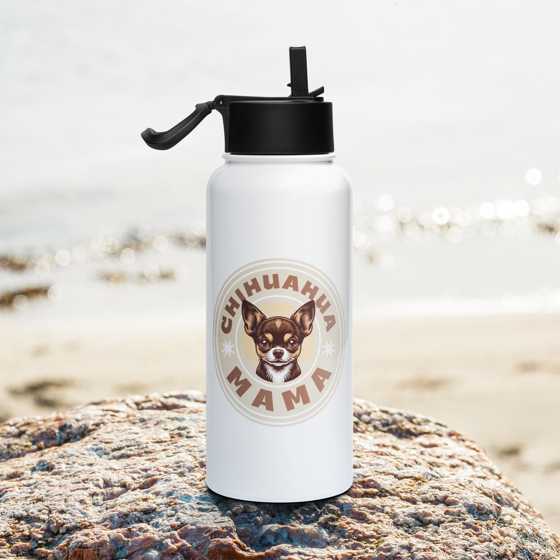 A white insulated water bottle with a black flip-straw lid stands on a large rock with a blurred beach background. The bottle features a circular graphic with the text 'CHIHUAHUA MAMA' and an illustration of a Chihuahua dog.